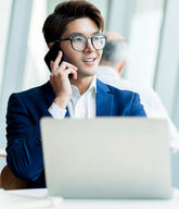 Man in a suit talking on a phone with a laptop and coffee cup in front of him