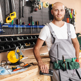 Man in a workshop wearing overalls with tools, standing in front of a workbench.