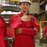 Person in red work uniform and white hard hat giving a thumbs-up in an industrial setting