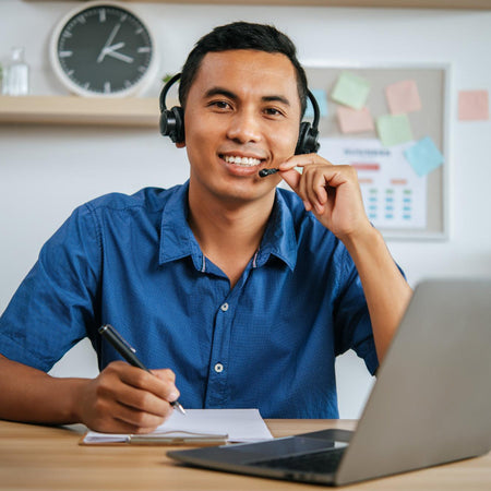 Man in a blue shirt with a headset sitting at a desk with a laptop and books.