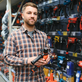 Man holding a power drill in a hardware store aisle with tools on shelves.