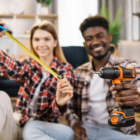 Two people in a living room with a woman holding a tape measure and a man holding a power drill.