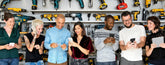 Group of people using digital devices in a workshop setting with tools on shelves.